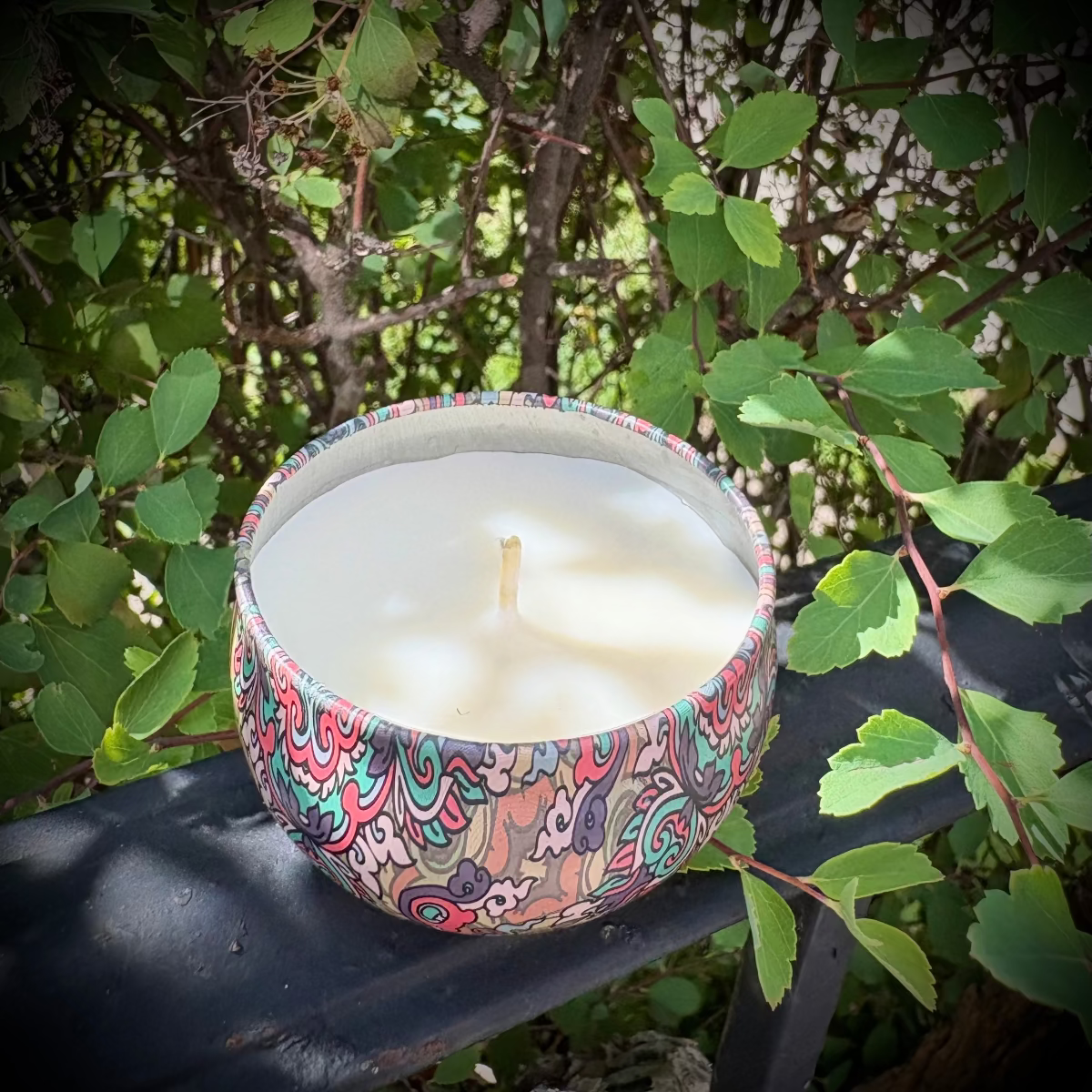 Decorative candle with a colorful pattern on a metal surface surrounded by green leaves.