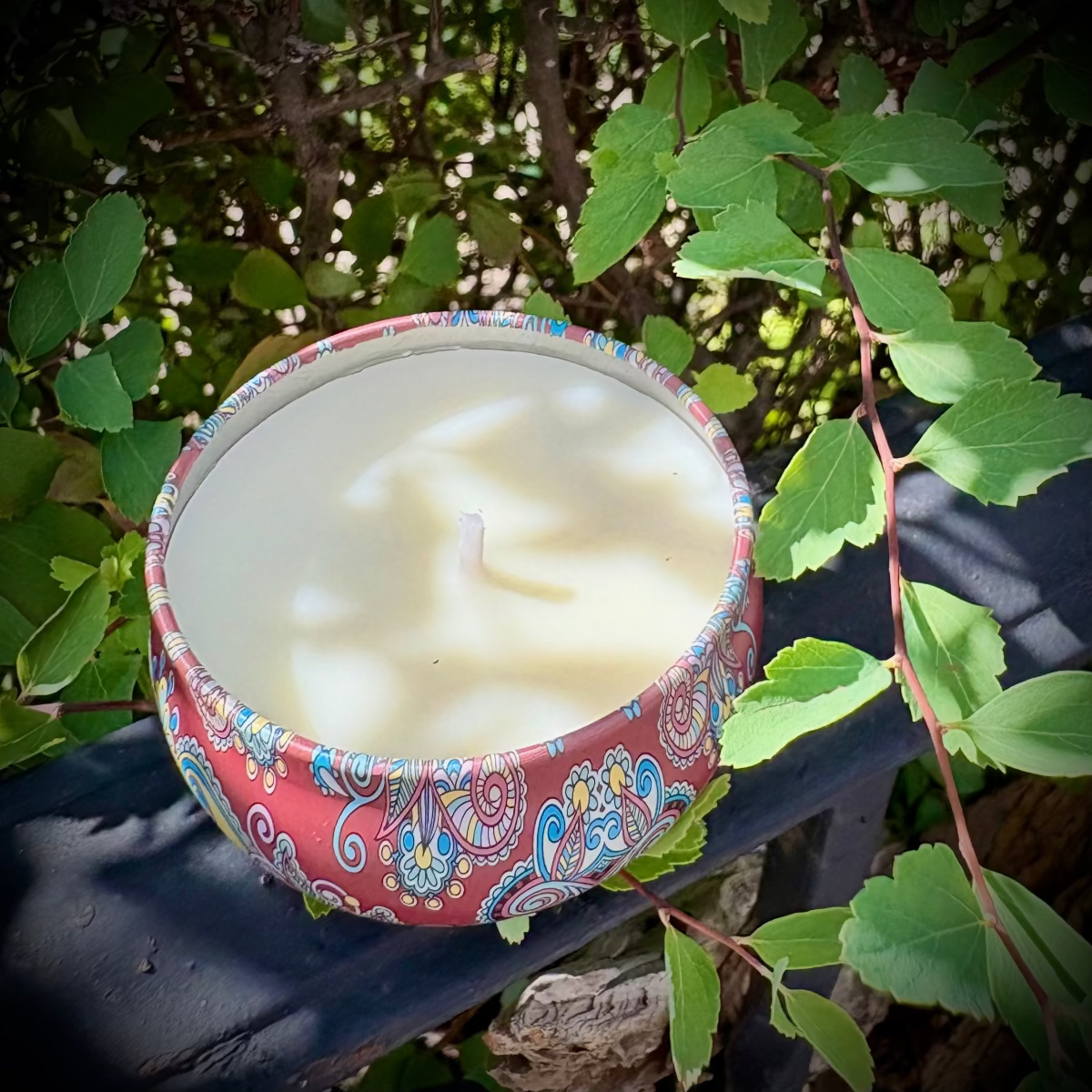 Candle in a colorful container surrounded by green leaves